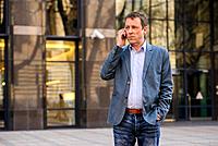 A middle age businessman standing in front of an office building while talking on his phone.