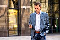 A middle age businessman standing in front of an office building while using his smartphone.