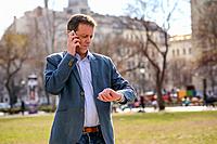 A middle age businessman standing in a park while talking on his phone and checking the time on his watch.