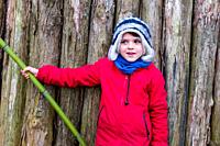 portrait of smiling 10 year old boy dressed in red jacket, leaning on a bamboo, with wooden planks background.