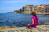 A preteen all dress in pink playing on the side of the bay of Saint Tropez. We can see the city village of Saint Tropez in France behind her.