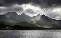 White house under Blaven mountains of Black Cuillin Hills with sun rays under dark clouds at Loch Slapin Isle of Skye Scotland UK.