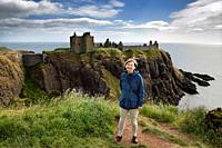 Scottish female tourist visitor at top of cliff above Old Hall Bay with Donnottar Castle ruins on the North Sea Scotland UK.