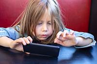 Portrait of five years old child with blue denim jacket, sitting in red sofa and black table in a restaurant, touching with finger a smartphone mobile...