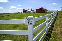 Sign on a fence, with a farm in the background, reading "All dogs must be on leash at all times".