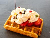 Chocolate syrup falling on a Belgian waffle with cream and colorful chocolates on pieces of strawberry on a slate plate.
