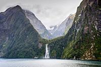 Ship near waterfall at Milford Sound Fjord in rainy weather and dramatic sky - South Island of New Zealand. This fiord is considered as one of the mos...
