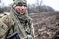 Portrait of soldier, modern combatant with dirty face, firearm replica, wearing camouflage uniform, beanie and masking cape on neck, standing on field...