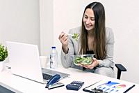 Smiling young woman sitting and eating a salad in coworking space. Horizontal indoors shot.