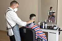 hairdresser grooming beard at barber shop to a client, both wear masks due to the corona virus pandemic.