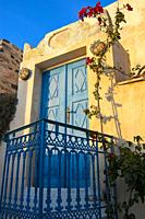 blue door in the wall of a typical house in santorini with pink flower plant.