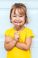 Adorable blond-haired girl eating ice cream, wearing a yellow shirt leaning against white background.