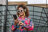 From below of happy stylish African American lady in earrings and sunglasses browsing internet on mobile phone while standing near building in dayligh...