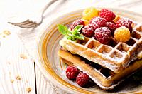 Belgian waffles served with raspberries and mint leaf dusted with powdered sugar on white wooden kitchen table.