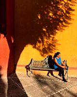 A woman sits in a street bench by the shadow of a tree in front of an orange wall in Peña de Bernal, Queretaro, Mexico.