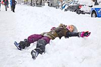 Happy seven years old child with coat, winter knitted hat, gloves and face mask removed lying in snow, laughing with open arms, in street of Madrid ci...