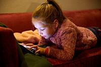 seven years old girl lying on red sofa at night touching digital screen mobile phone.