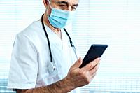 Mature male doctor - nurse wearing face mask, looking at mobile phone next to a hospital window. Covid-19 and medicine concept.