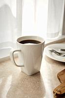 A white mug of coffee sitting on a counter of a sun streaked kitchen.
