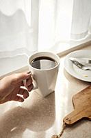 A woman's hand holding a white mug with coffee in a sunstreaked kitchen.