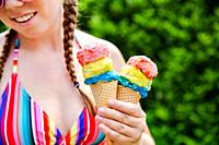 Beautiful girl holding two colorful ice cream with sprinkles wearing a rainbow colored swimsuit in the summer, pink sunglasses and braids happy vacati...