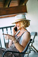 Portrait of a mature caucasian blonde young woman in her 50s talking on a mobile phone smiling and wearing a hat and sitting in a relaxed pose in a ro...