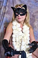 Woman wearing a faux fur boa, a black mask, black gloves and holding a whip sitting on a chair in a studio during a boudoir photo session.