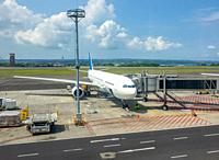 Sunny day at the Indonesian airport. An aircraft with an attached jet bridge for boarding and disembarking passengers. Ocean and jungle in the backgro...