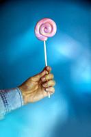 Colorful lollipop swirl on stick. Striped spiral multicolored pink candy on blue background, holding in hand sweet hard candy.