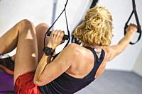 Real young mature caucasian woman in a sport wear at gym training arms exercises with trx fitness straps. Fitness concept.