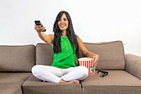 Optimistic Hispanic woman with bucket of popcorn smiling and switching channels on TV while sitting cross legged on couch during showtime at home.