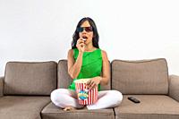 Hispanic female in casual clothes and 3D glasses eating fresh popcorn and watching film on TV while sitting cross legged on sofa in daytime at home.