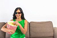 Glad Hispanic female in 3D glasses taking popcorn from bucket and smiling while resting on sofa and watching movie on weekend day at home.