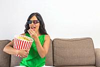 Young Hispanic woman eating crunchy popcorn from bucket while sitting on couch and watching 3D movie on weekend day in living room at home.