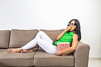 Side view of barefoot Hispanic female in casual clothes and 3D glasses eating fresh popcorn and watching interesting movie while chilling on couch in ...