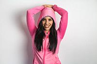 Cheerful Hispanic female model in hood of pink sports jacket keeping hands on head in studio on white background and looking at camera.