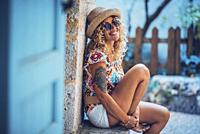 Portrait of beautiful smiling hipster young woman in sunglasses and straw hat sitting outdoors. Stylish tattooed woman in good mood spending leisure t...