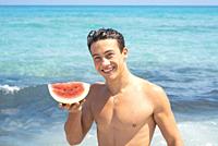 Young fit body boy portrait holding red fresh watermelon with blue ocean water in background. Summer holiday vacation season concept lifestyle people.