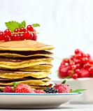 Stack of baked pancakes with fruits in a round plate on a white table.