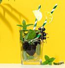 Transparent glass with lemonade, mint leaves, lemon slices and blackberries in the middle. Yellow background.