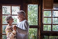 Old man and woman happy couple dancing at home in living room with outdoors background and windows. Senior people enjoy chalet for winter vacation tog...