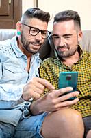 Happy young gay couple using mobile phone while sitting on a sofa in the living room. High quality photography.