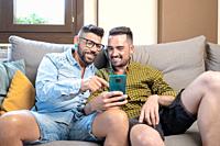Happy young gay couple using mobile phone while sitting on a sofa in the living room. High quality photography.