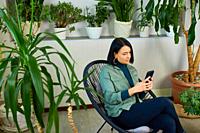 Woman chatting using smartphone, sitting on armchair at home, types message with happy smile, Concept of home garden, Biophilia design.
