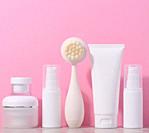 White plastic tubes and jars of cream, and a massage brush for facial cleansing on a pink background, items for cosmetic procedures.