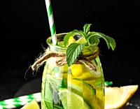 Lemonade in a transparent glass with lemon, lime, rosemary sprigs and mint leaves on a black background.