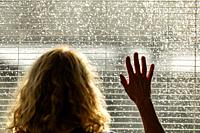 A woman looks through a bright window with blinds during a summer rain storm with sun.