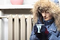 Middle age female model in parka and mittens hold coffee cup sitting on the floor under windowsill.