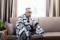 Middle age male model in hat and plaid hold coffee cup sitting on the sofa while using his smartphone.