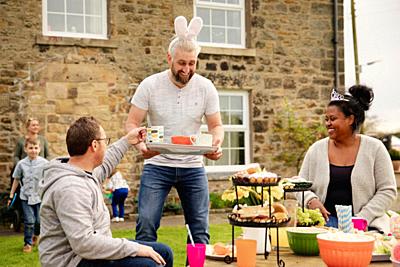 Mid adult man carrying a tray with cups of tea. He is handing out refreshments while wearing rabbit ears at a easter garden party.-stock-photo